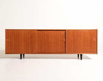 May include: A long, rectangular wooden sideboard featuring a warm, light brown wood grain finish. The unit has three sliding doors, each equipped with a small, square, integrated wooden handle. It is supported by four tapered black legs.