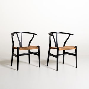Può includere: Due sedie in legno nero con schienali curvi e supporti a forma di Y. Ogni sedia presenta una seduta in corda intrecciata color marrone chiaro, quattro gambe dritte e una barra di supporto orizzontale che collega le gambe anteriori per stabilità.