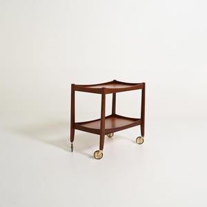 Op de afbeelding: Een houten serveerwagen met twee niveaus, een rijke bruine afwerking, gebogen randen aan beide rechthoekige planken en vier metalen zwenkwielen met goudkleurige accenten. De structuur wordt ondersteund door vier verticale houten poten.