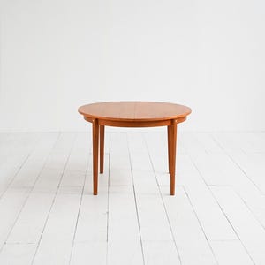 Puede incluir: Una mesa redonda de madera con un cálido acabado marrón rojizo. La mesa tiene cuatro patas delgadas y cónicas y un diseño simple y clásico. La mesa tiene aproximadamente 100 cm de diámetro.