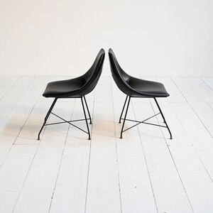 Peut inclure: Deux chaises noires avec dossiers et assises incurvés. Les chaises ont de fins pieds en métal noir qui se croisent près de la base. Les chaises sont placées face à face sur un plancher en bois blanc.
