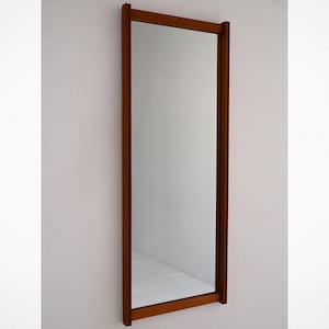 Puede incluir: Un espejo rectangular de cuerpo entero con marco de madera. El marco es de color marrón cálido y tiene un diseño simple y minimalista. El espejo está montado en una pared blanca, reflejando la habitación.