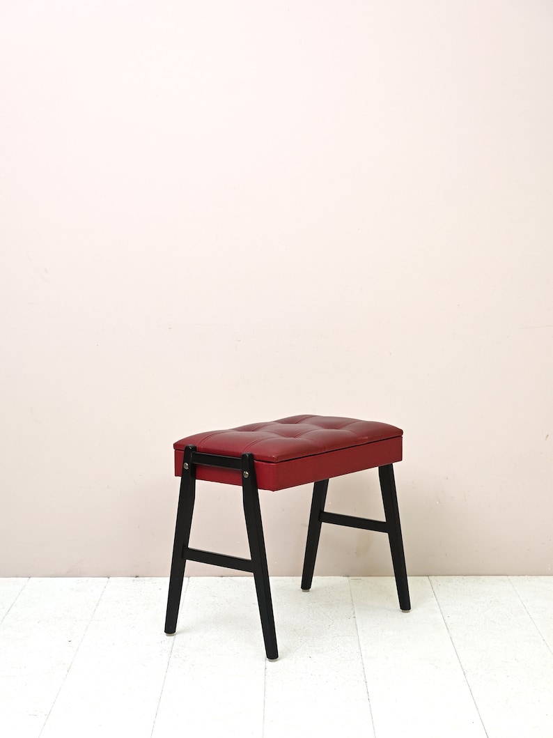 May include: A red upholstered piano bench with black wooden legs. The bench has a tufted top and a hinged lid.