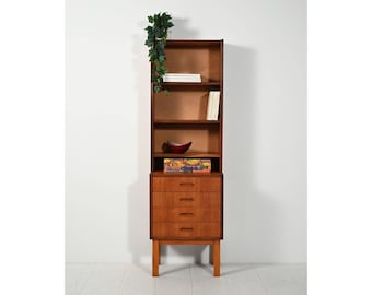 Puede incluir: Librería alta de madera de mediados de siglo con cuatro estantes y tres cajones. La librería es de color marrón cálido y tiene forma rectangular. Una pequeña planta decorativa cuelga de la parte superior. Libros y objetos decorativos en los estantes.