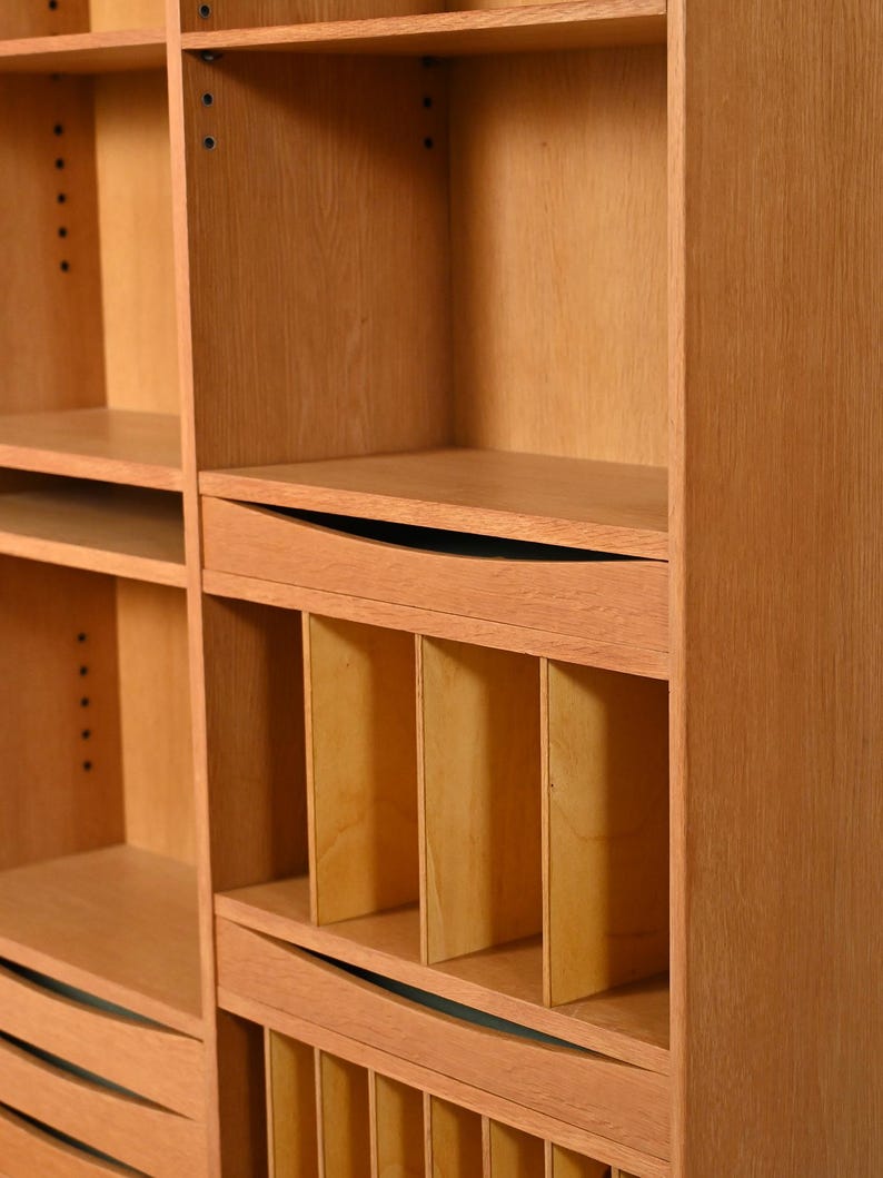 May include: A light brown wooden bookcase with multiple shelves and compartments. The bookcase has a natural wood grain finish and a simple, modern design. The shelves are of varying sizes, and some have curved edges. The bookcase is likely used for storage and display.