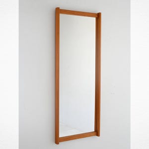 May include: A full-length mirror with a light brown wooden frame. The mirror is mounted on a white wall. The frame is approximately 5 cm thick and has a simple, minimalist design.