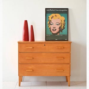 Op de afbeelding: Een licht houten dressoir met drie lades en een sleutelgat. Twee rode keramische vazen staan op het dressoir. Een zwart-wit poster met de tekst "Andy Warhol. Una retrospectiva" hangt aan de muur achter het dressoir.