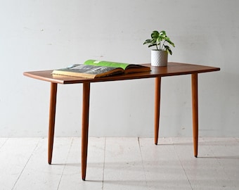 May include: A light wood coffee table with a rectangular top and four slender legs. A book and a potted plant are on the table.