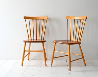 Könnte beinhalten: Zwei Holzstühle mit hellbraunem Finish. Jeder Stuhl hat eine Spindelrückenlehne und eine gebogene obere Schiene. Die Sitze sind dunkler braun. Die Stühle stehen vor einer weißen Wand und einem weißen Holzboden.