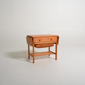 May include: A light brown wooden sewing table featuring a rectangular top with two drop-leaf sides, a single drawer with two small round knobs, a woven basket compartment underneath, and a slatted lower shelf supported by four straight legs.