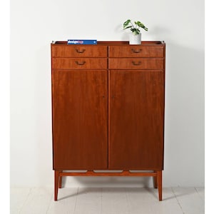 Può includere: Un mobile alto in legno con una calda finitura marrone-rossastra. Presenta due serie di cassetti e due ante. Una piccola pianta in vaso bianco e un libro sono sopra. Il mobile poggia su gambe sottili e angolate.