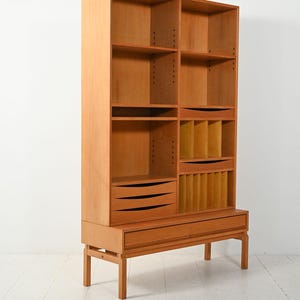 May include: A light brown wooden bookcase with multiple shelves, drawers, and compartments. The mid-century modern design features a rectangular shape with a base supported by four legs. The bookcase has a height of approximately 72 inches.