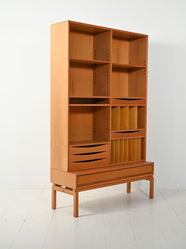 May include: A light brown wooden bookcase with multiple shelves, drawers, and compartments. The mid-century modern design features a rectangular shape with a base supported by four legs. The bookcase has a height of approximately 72 inches.