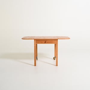 May include: A light brown wooden drop-leaf table featuring a central drawer with a circular knob. The table is supported by four straight legs, with two of the legs equipped with small metal casters for mobility, allowing the side leaves to fold down.