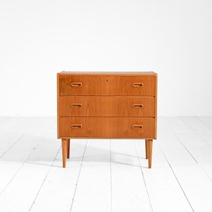 Könnte beinhalten: Ein dreischubladiger Holzschrank mit hellbraunem Finish. Der Schrank hat eine rechteckige Form mit drei Schubladen, jede mit einem eingelassenen Griff. Der Schrank steht auf vier konischen Beinen, und ein Schlüsselloch ist auf der obersten Schublade sichtbar.
