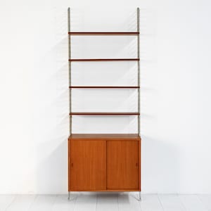 May include: A mid-century modern wall unit with a wooden cabinet base and open shelving. The cabinet has sliding doors and sits on metal legs. Above, five shelves are supported by metal rails, creating a stylish storage solution.