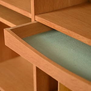 May include: A light brown wooden cabinet with an open drawer revealing a light green interior. The cabinet has multiple shelves and compartments, showcasing a minimalist design. The wood grain is visible, adding to the natural aesthetic.