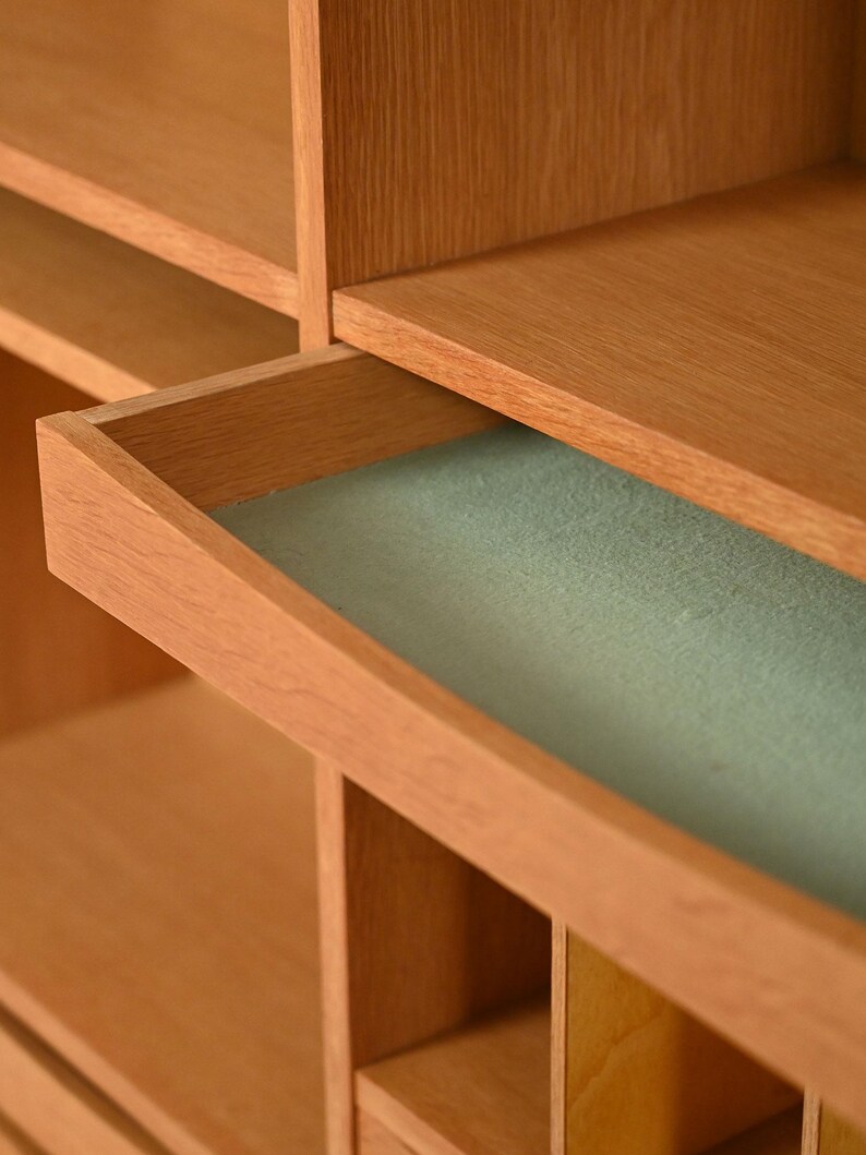 May include: A light brown wooden cabinet with an open drawer revealing a light green interior. The cabinet has multiple shelves and compartments, showcasing a minimalist design. The wood grain is visible, adding to the natural aesthetic.