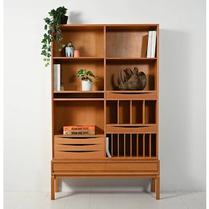 May include: A light brown wooden bookcase with multiple shelves, drawers, and compartments. The bookcase has a mid-century modern design and is filled with books, decorative items, and plants. The bookcase is against a white wall.
