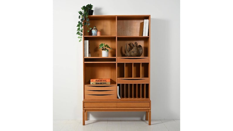 May include: A light brown wooden bookcase with multiple shelves, drawers, and compartments. The bookcase has a mid-century modern design and is filled with books, decorative items, and plants. The bookcase is against a white wall.