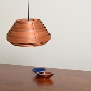 May include: A hanging pendant light fixture constructed from stacked, circular wooden slats in a warm brown tone, suspended by a black cord. Below, two small glass bowls are visible: one in a deep blue and one in a translucent amber color.