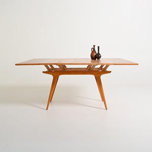 Puede incluir: Una mesa de comedor de madera de color marrón claro con un mecanismo de tablero extensible y cuatro patas cónicas. Dos pequeños jarrones decorativos, uno marrón y otro gris oscuro, están colocados sobre la superficie de la mesa.