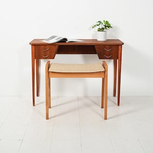 Könnte beinhalten: Ein Mid-Century-Modern-Schreibtisch aus Holz mit zwei Schubladen auf jeder Seite, ein passender Hocker mit gewebter Sitzfläche und eine kleine Topfpflanze. Auf dem Schreibtisch liegt eine offene Zeitschrift. Der Schreibtisch und der Hocker haben schlanke, sich verjüngende Beine.