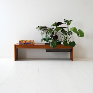 Puede incluir: Un banco de madera de estilo moderno de mediados de siglo con forma rectangular. El banco cuenta con una jardinera incorporada que contiene varias plantas verdes. Una caja colorida se encuentra en la superficie del banco. El banco está colocado contra una pared blanca y un suelo pintado de blanco.