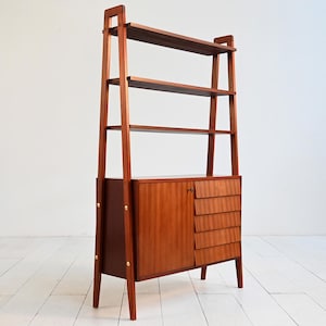 May include: A mid-century modern wooden bookcase with a warm, reddish-brown finish. It features open shelves, a cabinet with a door, and three drawers. The bookcase has a ladder-style frame and is set on angled legs.