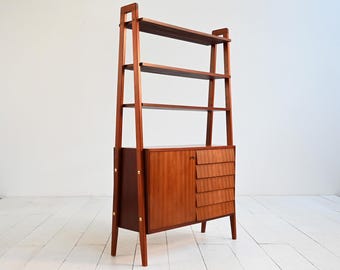 以下が含まれることがあります： 温かみのある赤茶色の仕上げが施された、ミッドセンチュリーモダン様式の木製本棚。 オープンシェルフ、扉付きのキャビネット、3つの引き出しを備えています。 本棚は梯子型のフレームで、角度の付いた脚の上に設置されています。