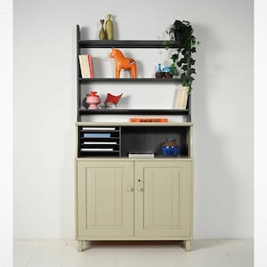 Puede incluir: Un aparador de dos puertas de color verde claro con una balda superior negra. Las baldas muestran objetos decorativos como jarrones, un caballo de madera y libros. El aparador tiene un organizador de cartas incorporado y un cuenco azul pequeño.