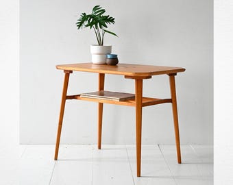 May include: A mid-century modern style wooden side table with a lower shelf. The table has a rectangular top and tapered legs. A potted plant and two small ceramic containers sit on top of the table. The table is made of light-colored wood.