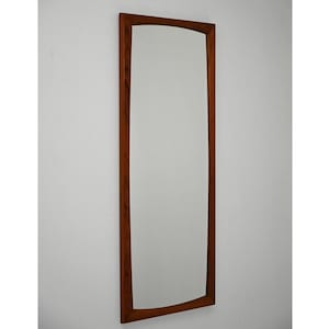 Puede incluir: Un espejo alto y rectangular con marco de madera. El marco es de color marrón cálido y tiene una parte superior e inferior ligeramente curvadas. El espejo está montado en una pared blanca.