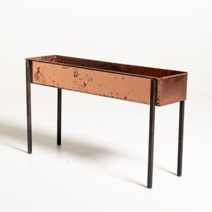 May include: A rectangular copper planter box with a weathered, oxidised finish, supported by four thin, dark metal legs. The planter features a long, narrow trough design with visible signs of ageing and patina on the exterior copper surfaces.