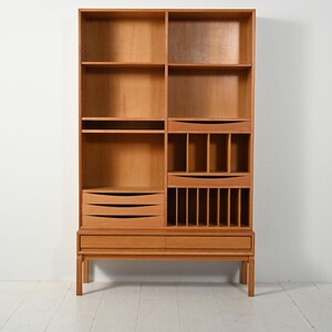 May include: A light brown wooden bookcase with multiple shelves, drawers, and compartments. The bookcase has a mid-century modern design with a rectangular shape and a raised base. The wood grain is visible throughout the piece.