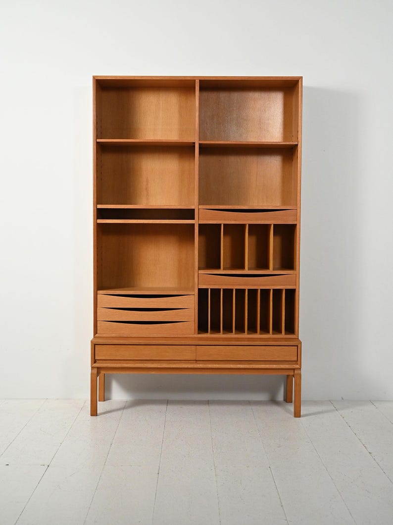 May include: A light brown wooden bookcase with multiple shelves, drawers, and compartments. The bookcase has a mid-century modern design with a rectangular shape and a raised base. The wood grain is visible throughout the piece.