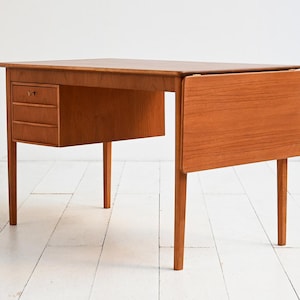 Könnte beinhalten: Ein Mid-Century-Schreibtisch aus Holz mit einer Klappverlängerung. Der Schreibtisch hat drei kleine Schubladen auf der linken Seite und eine rechteckige Tischplatte. Das Holz hat einen warmen, rötlich-braunen Farbton. Der Schreibtisch ist ca. 120 cm lang.