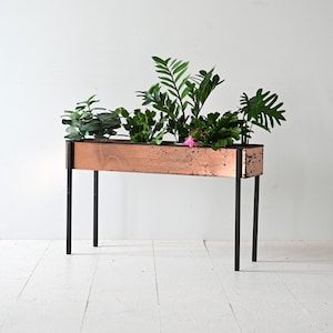Può includere: Un vaso da fiori in rame con gambe in metallo nero, riempito di piante verdi e fiori rosa. Il vaso è su un pavimento di legno bianco.