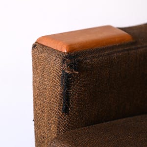 Pu&ograve; includere: Una poltrona in tessuto marrone con una parte superiore in legno. Il tessuto ha una trama tessuta e un piccolo strappo vicino alla parte superiore.