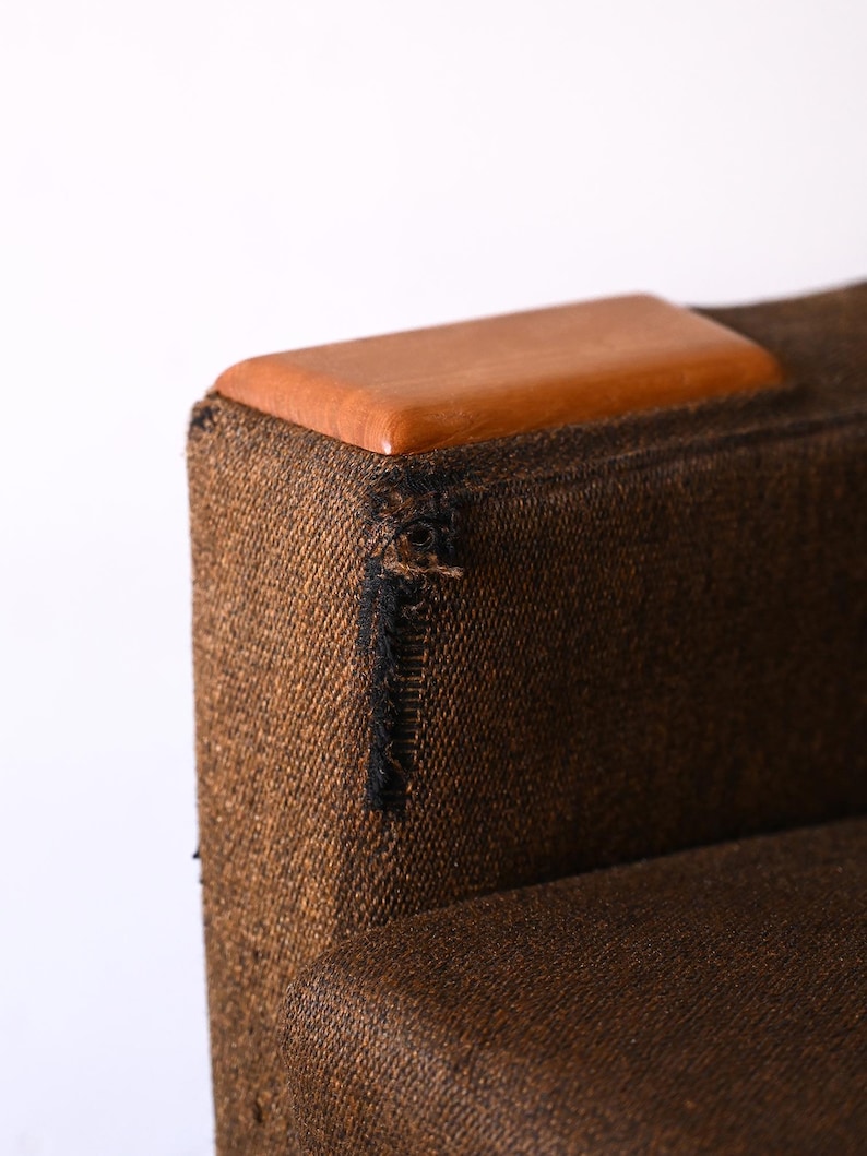 Pu&ograve; includere: Una poltrona in tessuto marrone con una parte superiore in legno. Il tessuto ha una trama tessuta e un piccolo strappo vicino alla parte superiore.