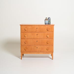 Puede incluir: Una cómoda de madera de color marrón claro con cuatro cajones horizontales, cada uno con dos pomos redondos de madera y una cerradura central. Dos pequeños jarrones decorativos de cerámica con patrones azules y verdes descansan encima.