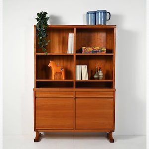 Puede incluir: Un mueble de madera de mediados de siglo con estanterías abiertas y dos puertas inferiores. El mueble es de color marrón cálido y presenta una figura de caballo naranja, libros y tazas azules. Una planta verde cuelga de la balda superior.
