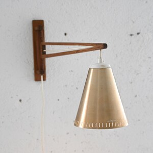 May include: A wall-mounted light fixture with a wooden arm and a gold-colored cone-shaped shade. The shade has a white rim and a series of small holes around the bottom edge.