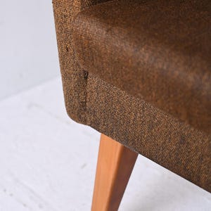 Pu&ograve; includere: Una poltrona in tessuto marrone con gambe in legno. La poltrona ha uno schienale imbottito e una seduta leggermente curva.