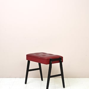 K&ouml;nnte beinhalten: Ein roter gepolsterter Hocker mit schwarzen Holzbeinen. Der Hocker hat eine gesteppte Oberseite und ein schlichtes Design.