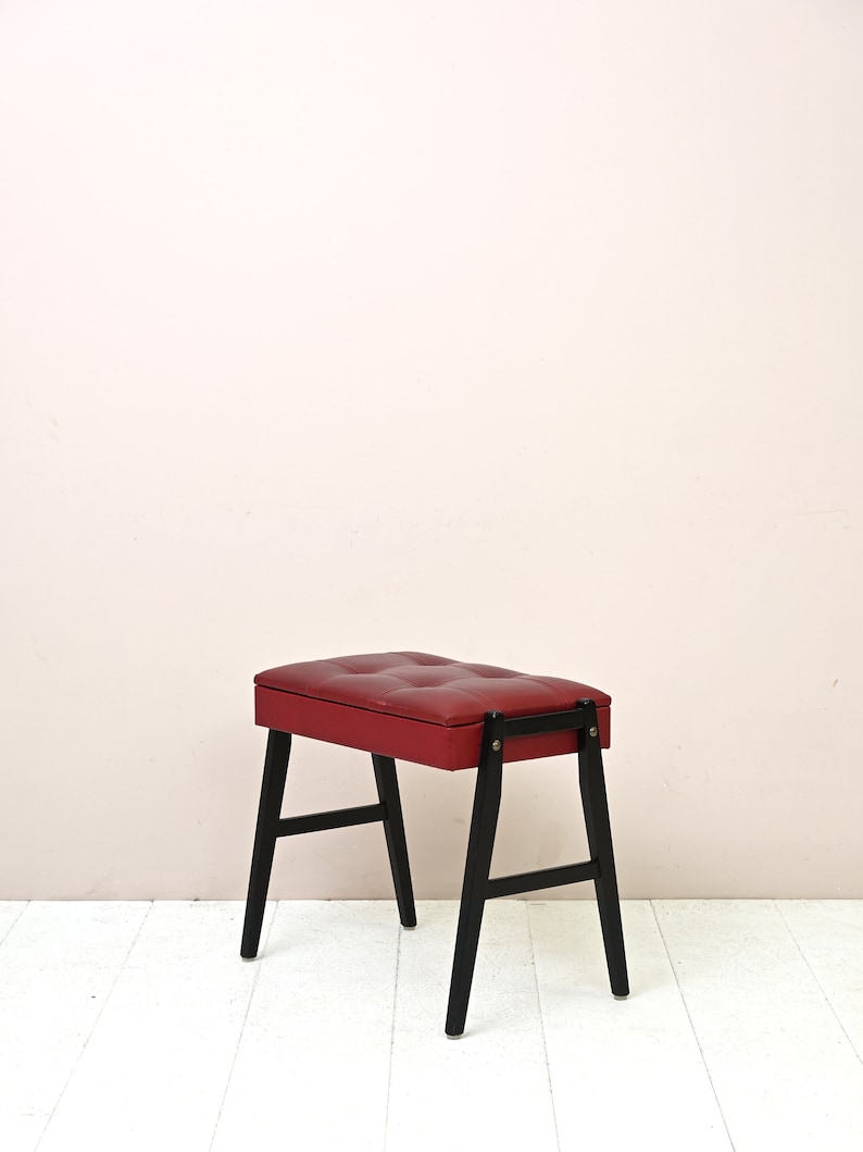 K&ouml;nnte beinhalten: Ein roter gepolsterter Hocker mit schwarzen Holzbeinen. Der Hocker hat eine gesteppte Oberseite und ein schlichtes Design.