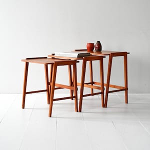 May include: A set of three wooden nesting tables in a row, each a different size. They have a warm, reddish-brown finish and a simple, mid-century modern design. A small, red ceramic pot and a decorative vase sit on the largest table. The tables are on a white floor.