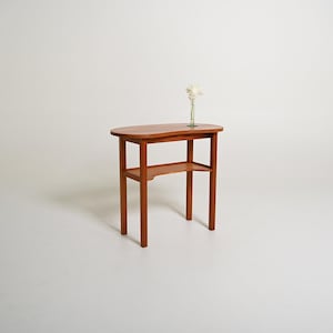 Peut inclure: Une console en bois de ton moyen dotée d'un plateau en forme de rein et d'une étagère inférieure soutenue par quatre pieds droits. Un vase soliflore en verre transparent contenant une fleur blanche est posé sur le côté droit du plateau.