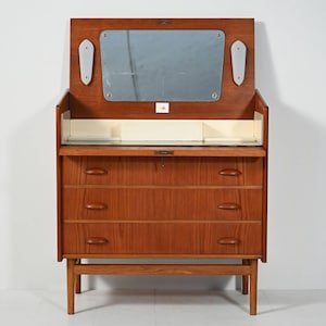 Puede incluir: Un tocador de madera de estilo moderno de mediados de siglo con espejo abatible y tres cajones. El tocador es de madera de tonos cálidos y presenta un espejo rectangular. El diseño incluye una pequeña balda y luces laterales decorativas.