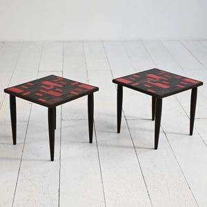 Può includere: Due tavolini quadrati con gambe nere e un piano scuro con un motivo geometrico rosso e nero. I tavoli sono in legno e hanno un design moderno della metà del secolo. I tavoli sono posizionati su un pavimento di legno bianco.
